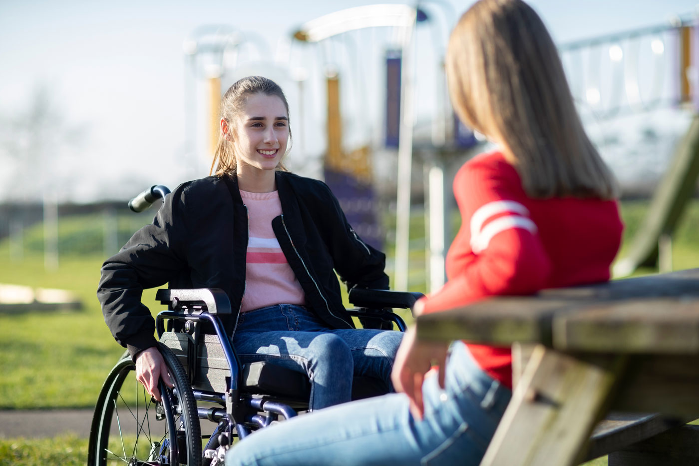 Two young friends chatting at outdoor bench, one is in a wheelchair